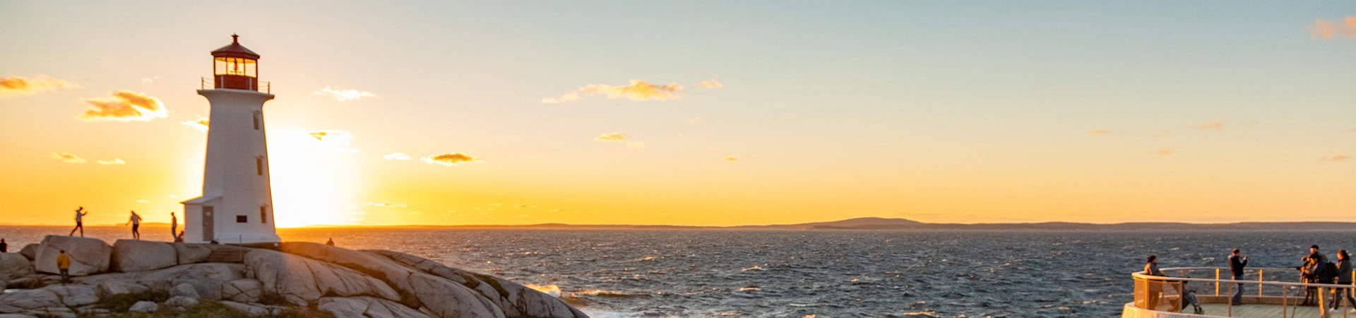 Ein weißer Leuchtturm mit roter Spitze steht links auf runden Klippen vor einem goldenen Sonnenuntergang über dem Meer, während rechts Menschen von einer Aussichtsplattform aus zuschauen.