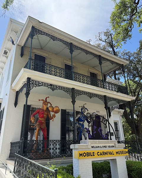 Die zweistöckige, historische Fassade des Mobile Carnival Museum im Bernstein-Bush-Haus zeigt kunstvolle, gusseiserne Balkone, zwei große Narrenfiguren auf der Veranda und ein Schild mit der Aufschrift „William & Emily Hearin Mobile Carnival Museum“ im Vordergrund unter blauem Himmel.