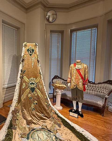 Eine prachtvolle, mit goldenen Stickereien und grünem Samt verzierte königliche Schleppe sowie ein passendes goldenes Kostüm mit roter Schärpe sind auf Schaufensterpuppen in einer historischen Zimmerausstellung des Mobile Carnival Museum in Mobile präsentiert.
