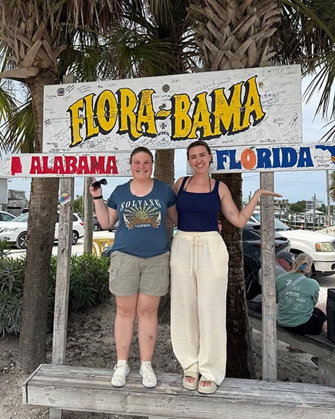 Zwei lächelnde junge Frauen stehen nebeneinander auf einer Holzbank vor dem berühmten Grenzschild der Flora-Bama Lounge, Package & Oyster Bar, das mit vielen Unterschriften versehen ist und die Aufschrift „FLORA-BAMA“ sowie darunter zwei Richtungspfeile nach „ALABAMA“ und „FLORIDA“ zeigt, umrahmt von zwei großen Palmenstämmen.
