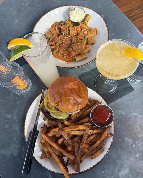 Ein saftiger Burger mit Käse, Salat, Tomaten und Zwiebeln wird zusammen mit einem großen Berg knuspriger, hausgemachter Pommes frites und einem Schälchen Ketchup auf einem Teller serviert, während daneben ein Teller mit Rigatoni-Pasta in einer reichhaltigen Fleisch- oder Tomatensauce sowie ein Glas Limonade und ein fruchtiger Cocktail auf dem Tisch stehen.