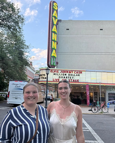 Zwei lächelnde junge Frauen stehen nebeneinander im Vordergrund einer Straße direkt vor dem historischen Savannah Theatre am Chippewa Square in Savannah, Georgia. Im Hintergrund ragt das große, vertikale Neon-Schild mit der grün leuchtenden Aufschrift „SAVANNAH“ empor, während die weiße Anzeigetafel (Marquee) in roten Buchstaben das Stück „ELVIS, JOHNNY CASH - MILLION DOLLAR QUARTET - LIVE ON STAGE“ ankündigt und links ein weißer Reisebus parkt.