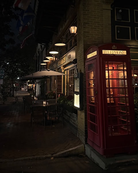 Eine ikonische, rot leuchtende britische Telefonzelle steht bei Nacht direkt an der Straßenecke vor der beleuchteten Fassade des bekannten Six Pence Pub in Savannah, Georgia, während auf dem schattigen Gehweg davor Tische und Stühle unter einem Sonnenschirm im warmen Licht der Außenlaternen angeordnet sind.