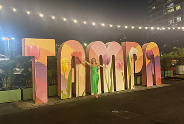 Eine Frau in einem leuchtend grünen Kleid steht lächelnd vor einem großen, bunten „TAMPA“-Schriftzug im Freien bei Nacht. Die riesigen Buchstaben sind mit farbenfrohen Tulpenmotiven gestaltet, und eine Kette aus warmen Lichtern zieht sich über die Szene im Hintergrund.
