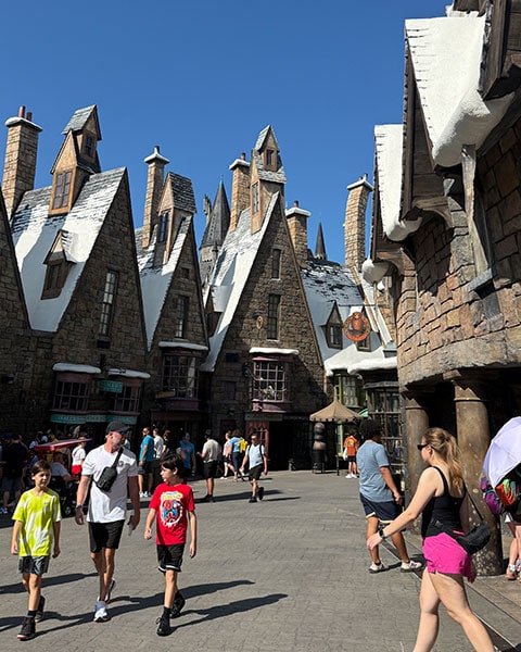 Menschen schlendern durch die schneebedeckten Steinhäuser von Hogsmeade in der Wizarding World of Harry Potter unter blauem Himmel.