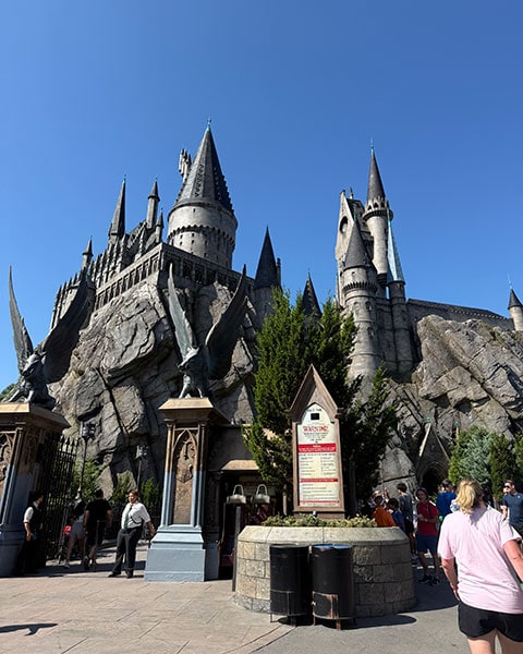 Das majestätische Schloss Hogwarts auf hohen Felsen mit zwei geflügelten Ebersäulen am Eingang der Attraktion „Harry Potter and the Forbidden Journey“ unter blauem Himmel.