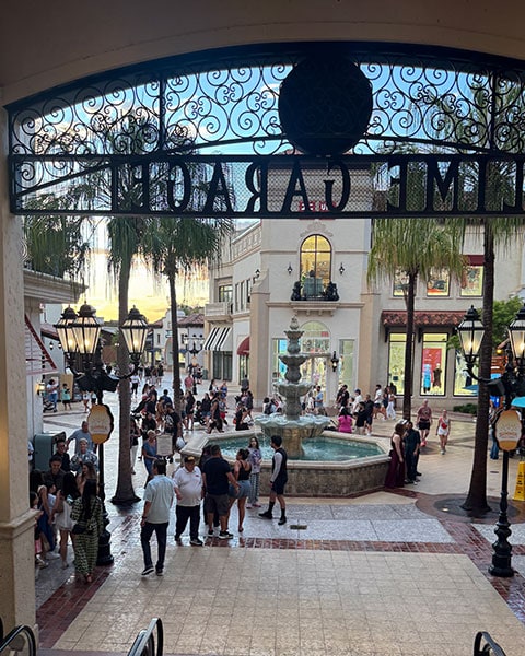 Menschen laufen an einem Steinbrunnen in Disney Springs vorbei, fotografiert unter dem schmiedeeisernen Bogen der „Lime Garage“.
