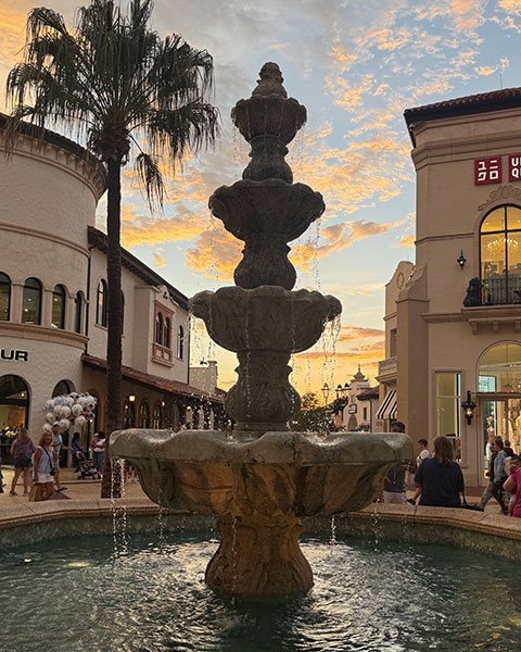 Ein mehrstöckiger Steinbrunnen plätschert im Zentrum von Disney Springs vor Palmen und Geschäften bei einem farbenfrohen Sonnenuntergang.
