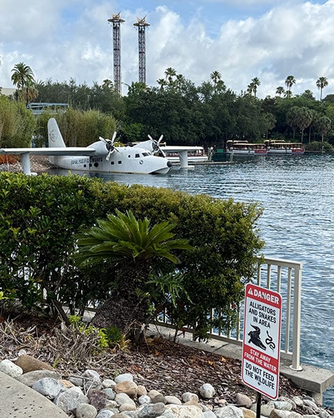 Ein weißes Propeller-Wasserflugzeug liegt am Ufer einer Lagune bei den Universal Studios, im Vordergrund warnt ein Schild vor Alligatoren und Schlangen.