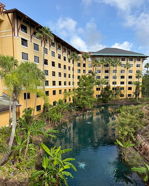 Ein gelber Hotelkomplex des Loews Royal Pacific Resort mit Palmen und Bananenstauden an einer angelegten Wasserstraße unter blauem Himmel.