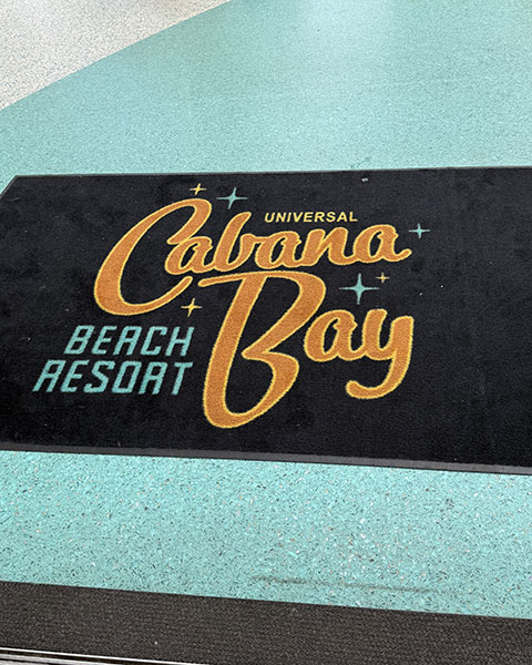 Eine schwarze Fußmatte mit dem gelb-türkisen Retro-Logo des „Universal Cabana Bay Beach Resort“.
