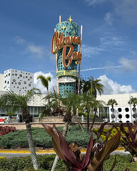 Das große, türkisfarbene „Cabana Bay Beach Resort“-Schild im Retro-Stil der 1950er Jahre steht umgeben von Palmen und tropischen Pflanzen vor blauem Himmel.