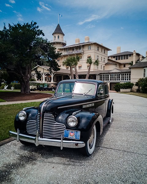 Ein glänzender, schwarzer Buick Oldtimer aus den späten 1930er Jahren steht im Vordergrund auf einer gepflasterten Auffahrt vor der prachtvollen, historischen Hotelanlage des Jekyll Island Club Resort auf Jekyll Island in Georgia, während im Hintergrund der markante weiße Aussichtsturm des Resorts mit einer blauen Flagge in den leicht bewölkten Himmel ragt.