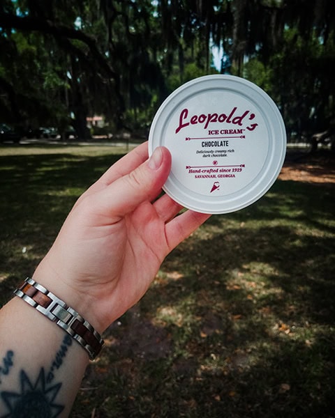 Eine Hand mit einem tätowierten Handgelenk hält einen weißen Eisbecher-Deckel von Leopold's Ice Cream mit der Aufschrift „CHOCOLATE“ im Vordergrund hoch, während sich im schattigen Hintergrund der grüne Rasen und die großen Eichen des historischen Forsyth Park in Savannah, Georgia, erstrecken.