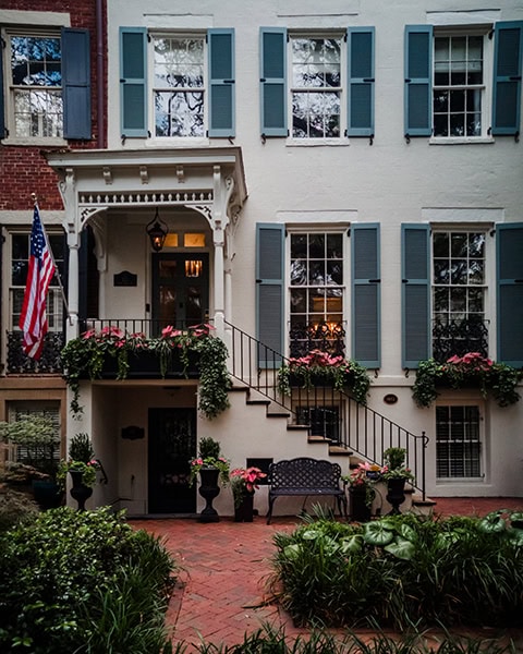Ein typisches, historisches Reihenhaus (Row House) im Landmark Historic District (nahe der berühmten Jones Street) in Savannah, Georgia, besticht durch eine weiß getünchte Backsteinfassade mit staubblauen Fensterläden. Eine geschwungene gusseiserne Freitreppe führt hinauf zum überdachten, weiß verzierten Eingangsbereich mit hängender Laterne und US-amerikanischer Flagge, während im Vordergrund ein rot gepflasterter Backsteinweg, üppiger Blumenschmuck und eine dekorative schwarze Metallbank zu sehen sind.