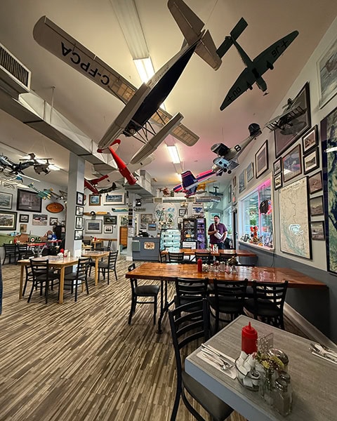 Blick in den gemütlichen Innenraum eines Cafés mit hellem Holzboden und Holztischen. An der Decke hängen zahlreiche große, detailreiche Modellflugzeuge, darunter ein silbernes Wasserflugzeug mit der Kennung „C-FPCA“. Die Wände sind dicht mit gerahmten historischen Fotos, Urkunden und einer großen Landkarte dekoriert.