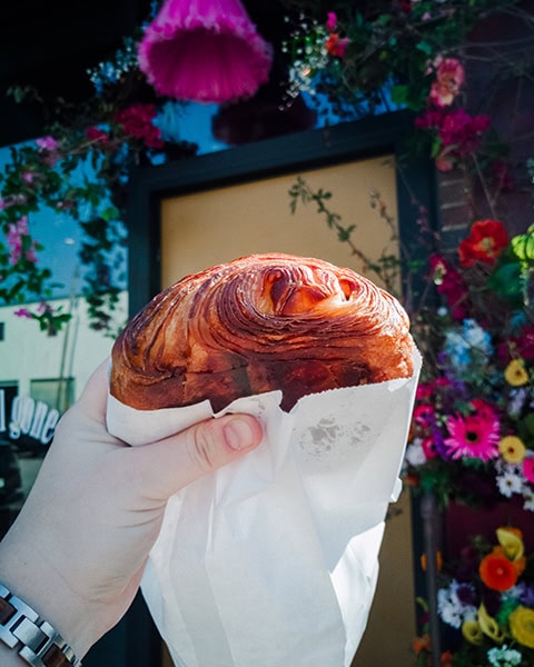 Eine Hand hält ein großes, goldbraun gebackenes und knusprig geschichtetes Croissant in einer weißen Papiertüte im Vordergrund, während im Hintergrund ein Eingang mit einer beigen Tür und üppigem Blumenschmuck zu sehen ist.