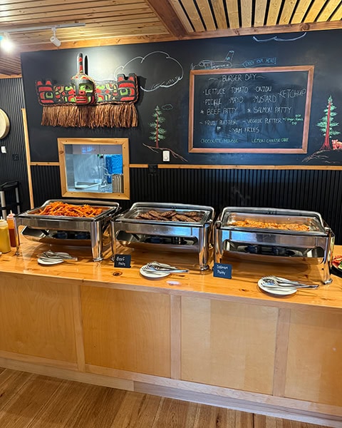 Ein Abendbuffet auf einer langen Holztheke mit drei großen Chafing Dishes aus Edelstahl. Auf kleinen Schildern davor steht „Beef Patty“ und „Salmon Patty“. An der schwarzen Wand dahinter hängt eine Kreidetafel mit dem Menü „Burger Day“ sowie eine traditionelle, rot-schwarze Holzmaske der First Nations.