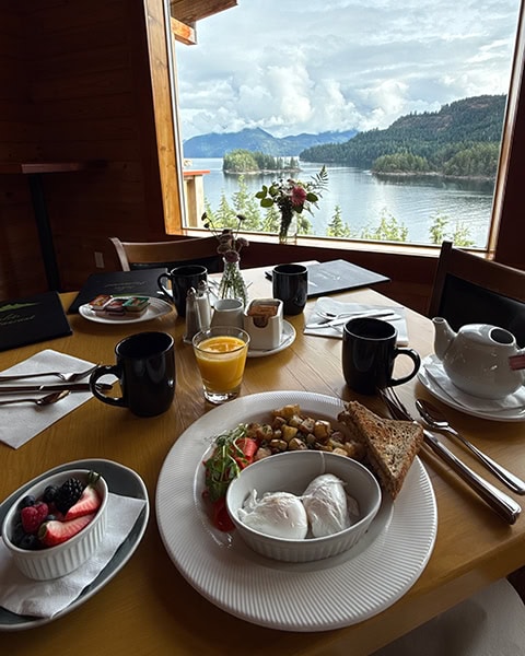 Ein reich gedeckter Frühstückstisch aus Holz steht vor einem großen Panoramafenster. Auf dem Tisch stehen ein Teller mit pochierten Eiern, Bratkartoffeln und Toast, eine Schale mit frischen Beeren, Saft und Kaffeetassen. Das Fenster rahmt eine weite Aussicht auf eine sonnige Bucht mit bewaldeten Inseln und Bergen unter Wolken ein.