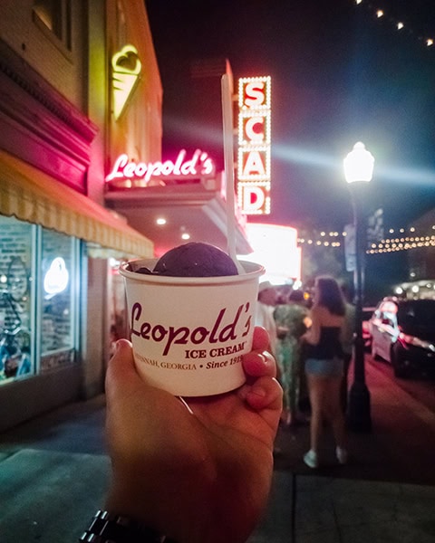 Eine Hand hält einen weißen Pappbecher mit einer Kugel dunklem Eis von Leopold's Ice Cream im Vordergrund, während im nächtlichen Hintergrund die rot leuchtenden Leuchtreklamen der traditionsreichen Eisdiele sowie das markante, vertikale Neon-Schild des SCAD Trustees Theater an der Broughton Street in Savannah, Georgia, zu sehen sind.