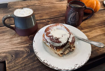 Eine große, frisch gebackene Zimtschnecke mit weißem Zuckerguss auf einem rustikalen Teller, platziert neben zwei gefüllten Kaffeetassen auf einem Holztisch.