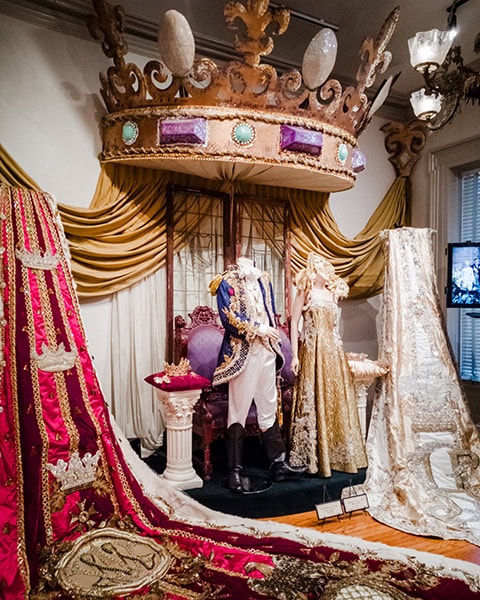 Eine riesige, kunstvoll verzierte goldene Krone mit lila und türkisfarbenen Schmucksteinen hängt als Baldachin über einer Museumsausstellung, die prunkvolle königliche Gewänder, Kleider und lange Schleppen auf Schaufensterpuppen vor goldenen Vorhängen zeigt.