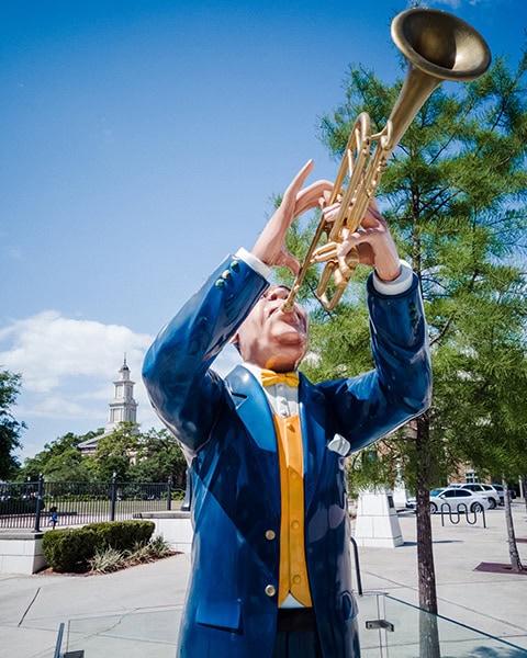 Eine lebensgroße, glänzende Skulptur eines Jazzmusikers im blauen Anzug und gelber Weste spielt eine goldene Trompete im Mardi Gras Park in Mobile, während im fernen Hintergrund das historische Gerichtsgebäude Mobile Government Plaza unter blauem Himmel zu sehen ist.