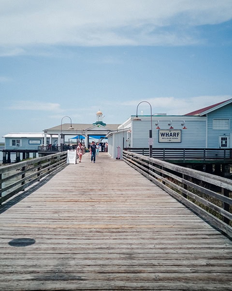 Ein langer Holzsteg mit hölzernen Geländern führt direkt auf das hellblaue Gebäude des Restaurants The Wharf auf Jekyll Island in Georgia zu, während im Hintergrund ein überdachter Pier-Bereich mit einem kleinen Turm und Passanten unter leicht bewölktem Himmel zu sehen sind.