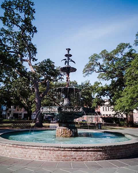 Der historische, gusseiserne Ketchum-Brunnen sprüht Wasser in einem großen, gemauerten Rundbecken im Zentrum des Bienville Square Parks, umgeben von schattigen, alten Eichen und Gehwegen unter blauem Himmel.