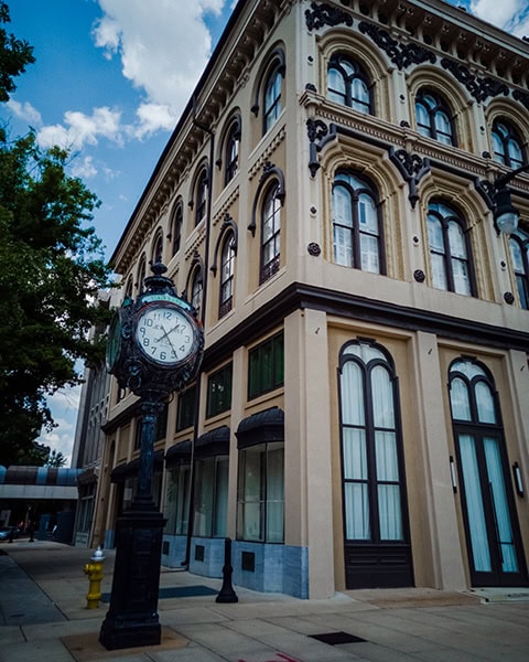 Eine freistehende, historische Standuhr aus schwarzem Gusseisen steht auf einem Gehweg vor einem mehrstöckigen, detailreich verzierten Eckgebäude mit Rundbogenfenstern unter blauem Himmel.