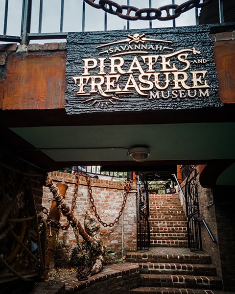 Ein großes, rustikales Holzschild mit der Aufschrift „SAVANNAH PIRATES AND TREASURE MUSEUM“ hängt über einem abwärts führenden Eingangsbereich im historischen Restaurant The Pirate's House in Savannah, Georgia, während darunter eine Backsteintreppe nach oben führt, die von schweren Eisenketten, Tauen und maritimer Dekoration gesäumt wird.