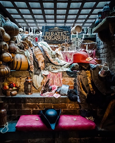 Eine detailreiche, düstere Ausstellungsnische zeigt ein Schild mit der Aufschrift „SAVANNAH PIRATES AND TREASURE MUSEUM“, umgeben von Skeletten in historischen Piratenkostümen, Holzfässern, alten Krügen, Kerzenleuchtern und einem roten Dreispitz-Hut auf einer gepolsterten Bank im Vordergrund unter einer gitterartigen Deckenbeleuchtung.