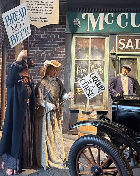 Eine detailreiche historische Szene im American Prohibition Museum in Savannah, Georgia, zeigt zwei lebensechte Schaufensterpuppen von Frauen in traditioneller Kleidung des frühen 20. Jahrhunderts, die während einer Demonstration Protestschilder mit den Aufschriften „BREAD NOT BEER“ und „LIQUOR IS A CURSE“ hochhalten. Im Hintergrund ist die nachgestellte Fassade des „McCarthy Saloon“ zu sehen, vor dem ein Mann neben einem historischen Ford Model T Oldtimer steht, während an der Backsteinwand darüber ein Zitat von Abraham Lincoln angebracht ist