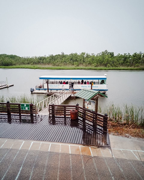 Das Ausflugsboot „Delta Explorer“ liegt an der Anlegestelle des 5 West Delta Safaris (nahe dem Meaher State Park) im Mobile-Tensaw River Delta, während eine Rampe von einem nassen Holzsteg zum Boot führt und im Hintergrund dichte Sumpfwälder unter bewölktem Himmel zu sehen sind.