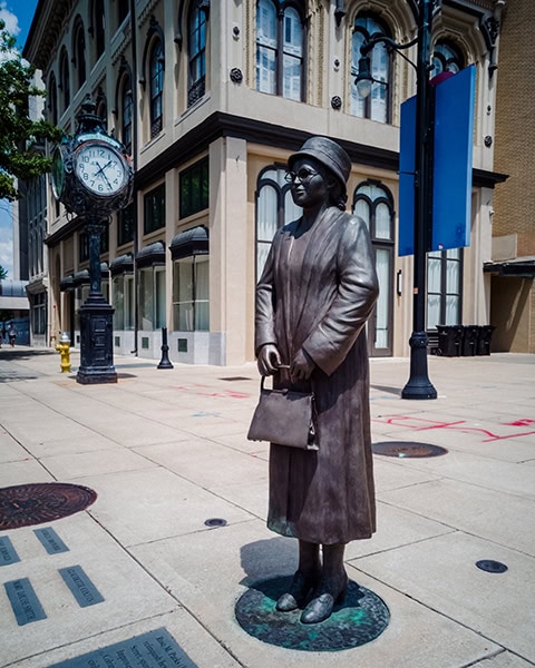 Eine lebensgroße Bronzestatue von Rosa Parks im Mantel mit Hut, Brille und einer Handtasche steht auf einem gepflasterten Gehweg vor einem historischen Eckgebäude, während im linken Hintergrund eine freistehende, historische Standuhr zu sehen ist.