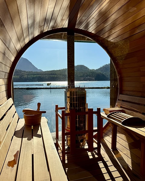 Blick aus dem Inneren einer hölzernen Fasssauna mit Holzbänken auf beiden Seiten und einem zentralen Saunaofen mit Schutzgitter. Ein riesiges, rundes Panoramafenster bietet eine freie Aussicht auf das sonnenbeschienene, glitzernde Fjordwasser und bewaldete Berge unter einem strahlend blauen Himmel.