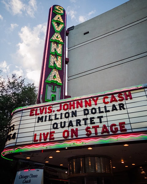 Ein großes, vertikales Neon-Schild mit der grün leuchtenden Aufschrift „SAVANNAH“ ragt an der Fassade des historischen Savannah Theatre in Savannah, Georgia, empor, während die weiße Anzeigetafel (Marquee) darunter in roten und schwarzen Buchstaben die Show „ELVIS, JOHNNY CASH - MILLION DOLLAR QUARTET - LIVE ON STAGE“ ankündigt.