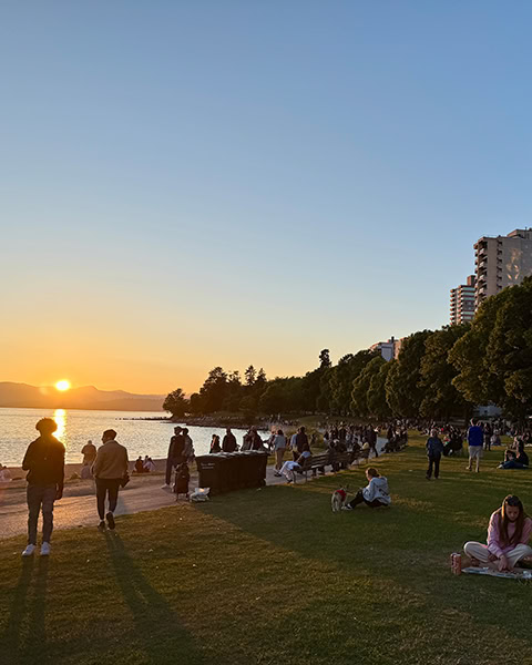 Menschen entspannen sich bei Sonnenuntergang auf einer Rasenfläche an einem Strand. Im Hintergrund sind das Meer, Berge am Horizont, Bäume und Hochhäuser zu sehen.