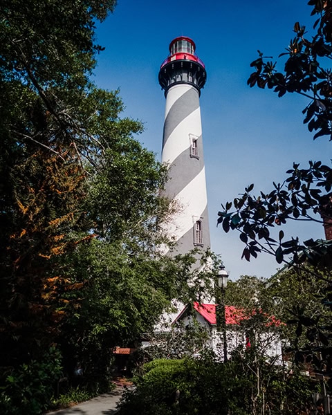 Der historische St. Augustine Lighthouse mit seinem markanten, schwarz-weiß spiralförmig gestreiften Turm und der leuchtend roten Laternenkappe ragt hinter dichten grünen Bäumen empor, während am Fuße des Turms ein weißes Wärterhaus mit rotem Dach steht und ein kleiner gepflasterter Fußweg im kühlen Schatten liegt.