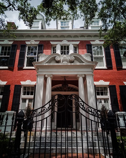 Die herrschaftliche Fassade des historischen Mills B. Lane House (auch bekannt als das Athenaeum des Ralston College) in Savannah, Georgia, beeindruckt durch ihr rotes Backsteinmauerwerk, weiße Fensterrahmen mit schwarzen Fensterläden und drei symmetrisch angeordnete Dachgauben. Ein prachtvoller, weißer Portikus im klassizistischen Stil mit korinthischen Säulen rahmt den Eingangsbereich ein, während im Vordergrund eine kunstvoll geschwungene, schwarze gusseiserne Zaunanlage den Blick auf die Stufen lenkt.