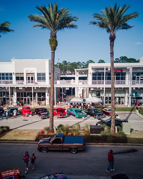 Mehrere klassische Autos und Hot Rods stehen während einer Autoshow aufgereiht auf der Main Street im Einkaufs- und Entertainmentkomplex The Wharf in Orange Beach, flankiert von zwei hohen Palmen im Vordergrund und einem modernen, zweistöckigen Geschäftsgebäude mit weißen Balkonen im Hintergrund unter blauem Himmel.