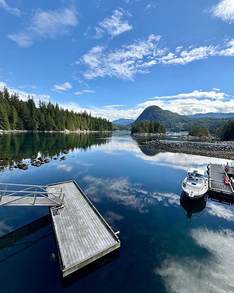Blick von oben auf eine spiegelglatte Bucht unter blauem Himmel mit weißen Wolken. Im klaren Wasser spiegeln sich der Himmel und die Wolken perfekt. Links führt eine Metallrampe zu einer hölzernen Schwimmplattform, rechts liegt ein weißes Motorboot an einem Steg. Im Hintergrund erstrecken sich dichte Nadelwälder und ein markanter runder Berg.