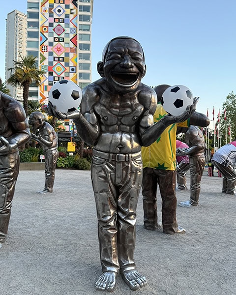Eine lebensgroße, glänzende Bronzeskulptur eines Mannes mit extrem lachendem Gesichtsausdruck hält zwei Basketbälle im Fußball-Design in den Händen. Im Hintergrund sind weitere Skulpturen der Gruppe sowie eine bunte Hausfassade zu sehen.