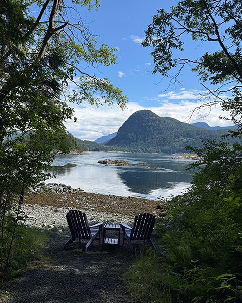 Blick durch eine Lücke in den Bäumen auf zwei dunkle Adirondack-Stühle mit einem kleinen Tisch dazwischen. Sie stehen an einem steinigen Strand vor einer ruhigen Bucht. Im Wasser spiegelt sich ein markanter, bewaldeter Berg unter einem blau-weißen Himmel.