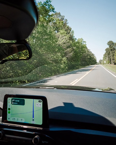 Der Blick aus dem Inneren eines Autos zeigt eine zweispurige Landstraße, die an dichten, grünen Bäumen vorbeiführt, während auf dem Navigationsbildschirm des Armaturenbretts eine Route angezeigt wird.