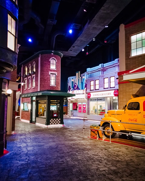 Eine nachgebaute, historische Kulissenstraße im Innenbereich des World of Coca-Cola Museums zeigt detailgetreue Häuserfassaden wie einen Lebensmittelladen namens „John’s Grocery“, ein Kioskgebäude und einen Oldtimer-Lieferwagen in leuchtendem Gelb auf gepflastertem Boden unter einer abgedunkelten Decke.