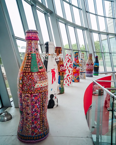 Eine Reihe von überlebensgroßen, kunstvoll und farbenfroh bemälten Coca-Cola-Flaschenskulpturen steht aufgereiht vor einer großen, geschwungenen Glasfassade in einem modernen Museumsinnenraum.