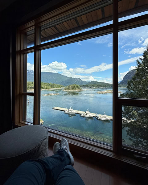 Blick aus der Ich-Perspektive eines Hotelzimmers. Auf dem Holzboden sind ausgestreckte Beine in dunklen Hosen und hellen Sneakern zu sehen. Ein riesiges Holzfenster rahmt den Ausblick auf eine ruhige Bucht mit einem langen Bootssteg ein, an dem drei weiße Motorboote liegen. Im Hintergrund erstrecken sich bewaldete Inseln und Berge unter blauem Himmel mit weißen Wolken.