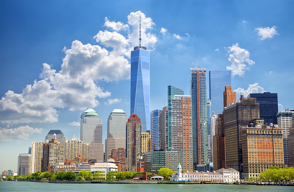 Die Skyline von Lower Manhattan in New York City mit dem One World Trade Center unter blauem Himmel mit weißen Wolken. Im Vordergrund stehen dichte Wolkenkratzer am Ufer des Hudson Rivers.